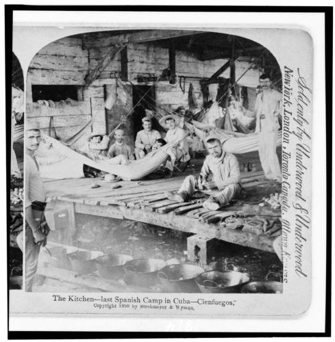 La Cocina- último campamento español en Cuba- CienfuegosResumenDos hombres cocinando mientras otros en un refugio de madera miran.1899