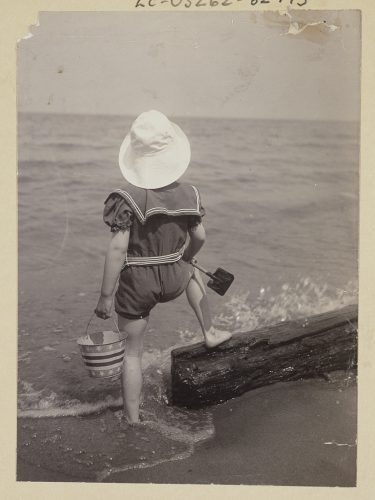 Un niño en verano (parte trasera)ResumenVista trasera de un niño en la playa, al borde del agua, sosteniendo un pequeño cubo y una pala de juguete. 